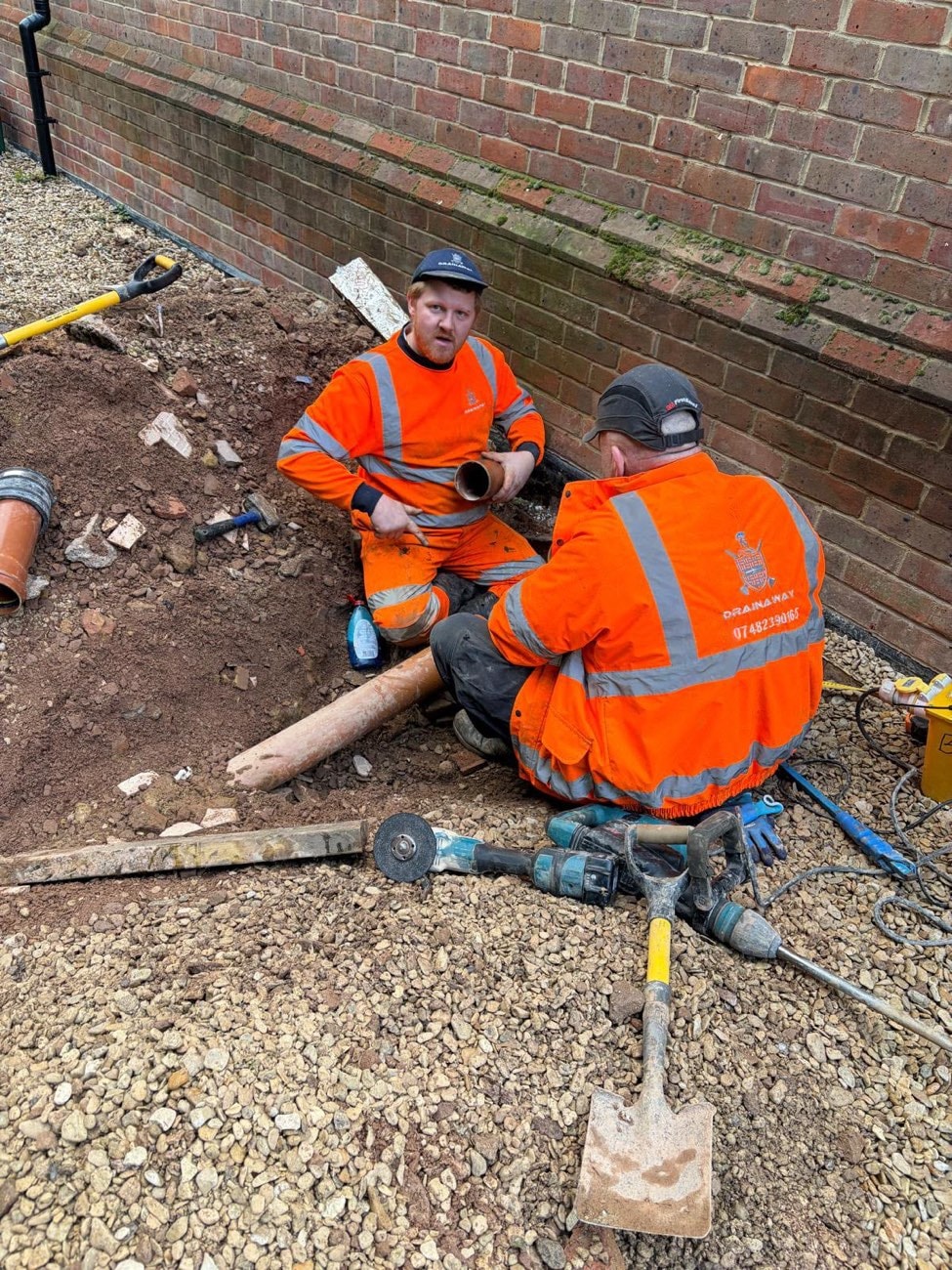 Two DrainAway engineers in orange high-visibility gear cutting pipe beside an excavation