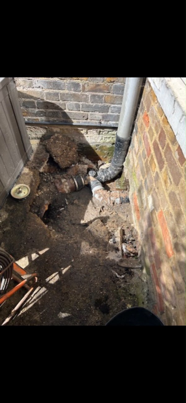Wet, contaminated ground at the base of a downpipe beside a brick wall, with the drain pipework exposed.