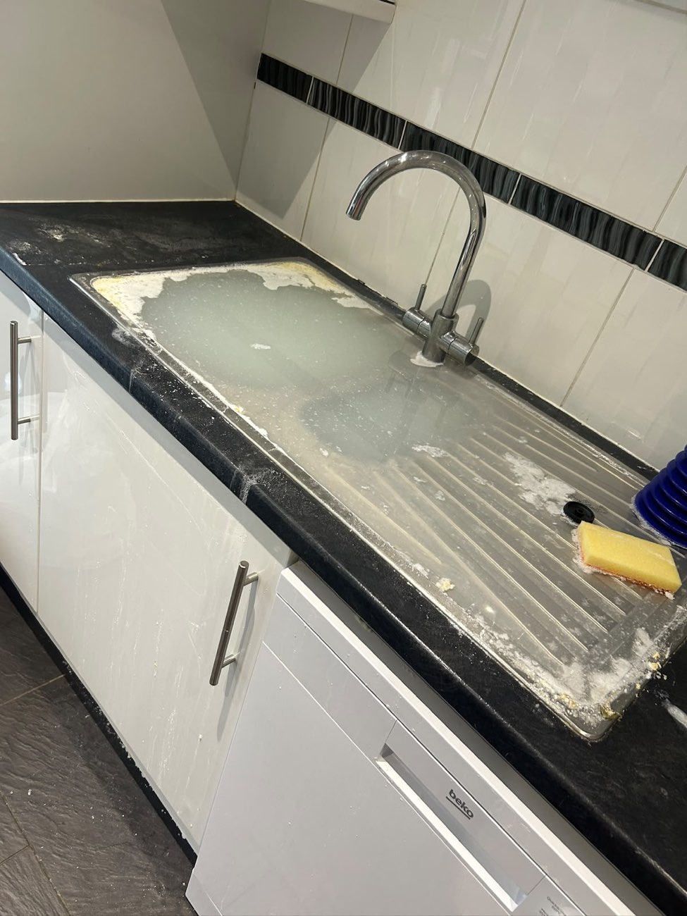 Kitchen sink overflowing with dirty standing water that has spilled across the black worktop and down the white units.