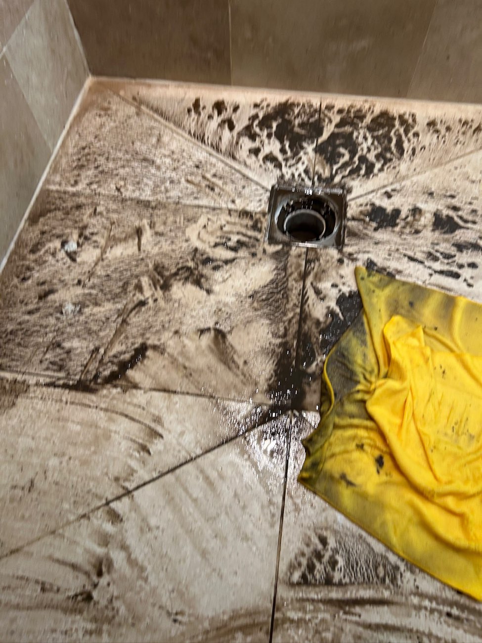 Tiled shower floor around a square chrome drain covered in dark silt and dirty water that has backed up out of the waste.