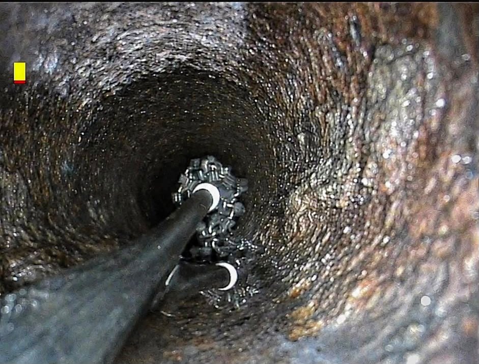 CCTV still of a rotating chain cutting head part way along a pipe caked in hard, dark scale.