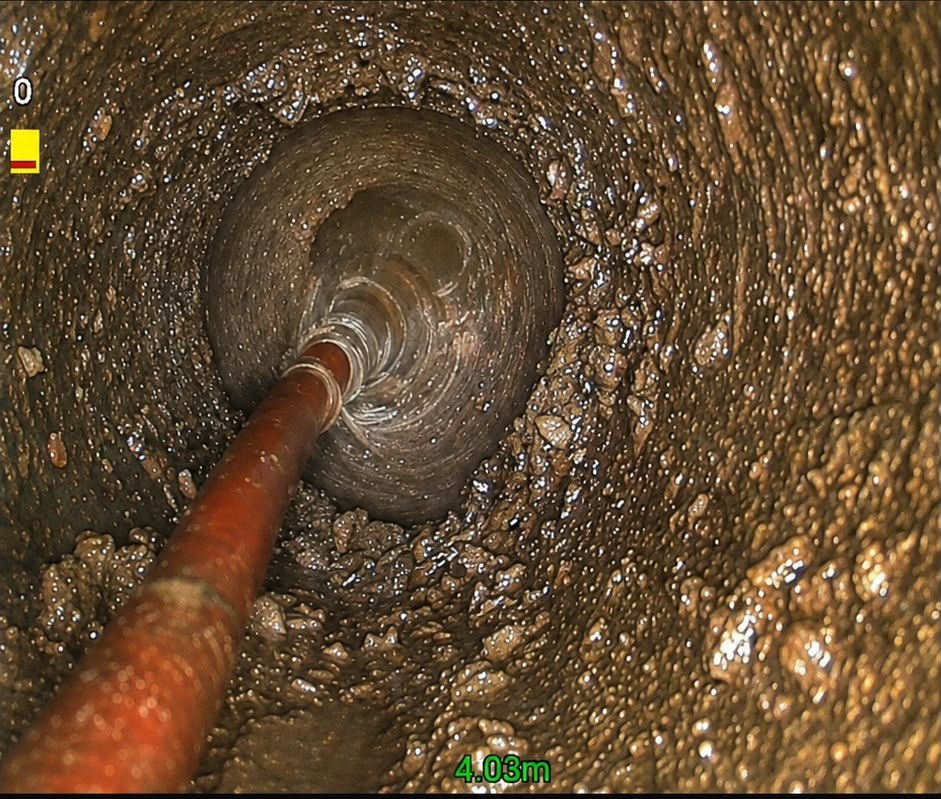 CCTV still of a descaling nozzle on a red hose four metres along a pipe heavily coated in hard brown build-up.