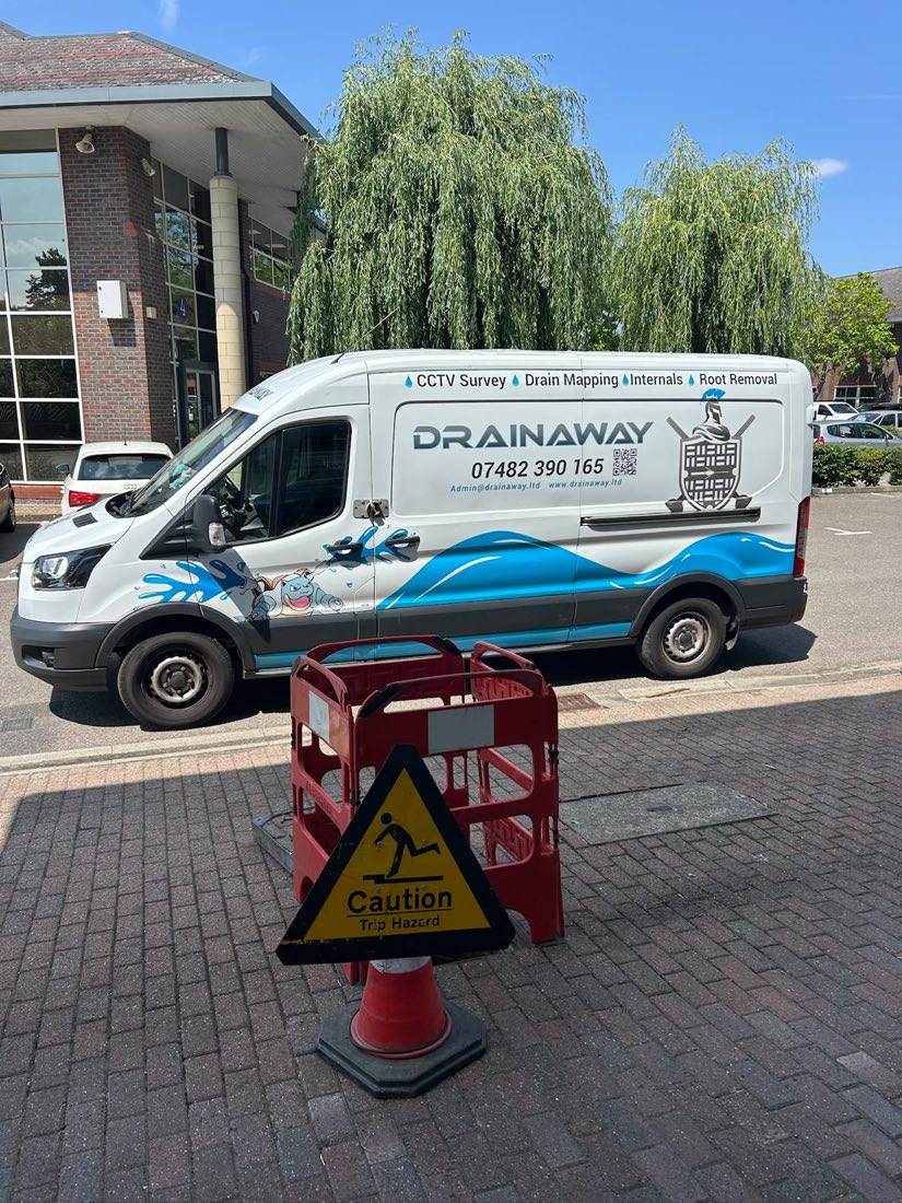 A liveried DrainAway van parked outside a modern commercial building with barriers and a caution sign set out.