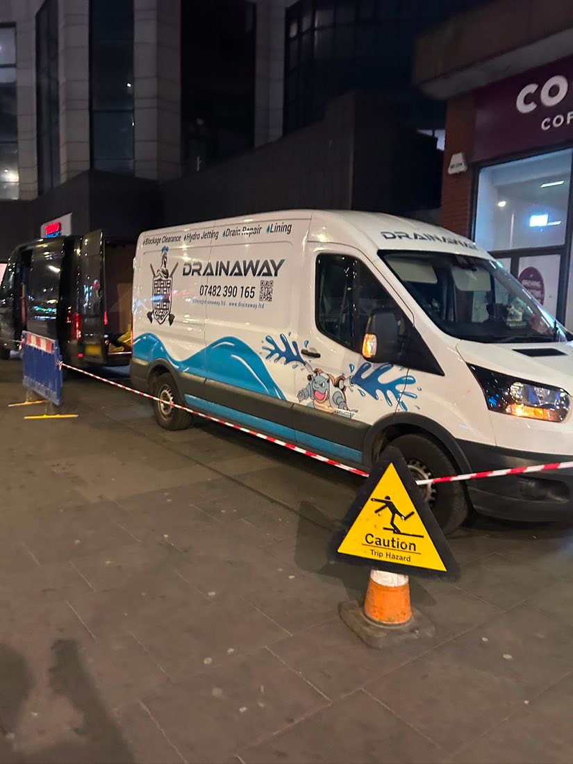 A liveried DrainAway van parked on a town street at night with barriers around the working area.